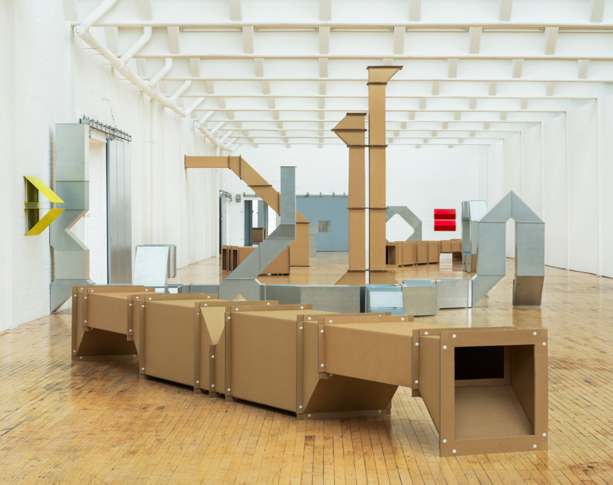 Installation of cardboard and tin-looking sculptures in large room with white walls and a wood floor.